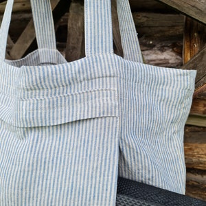 May include: A striped tote bag made of natural linen fabric with blue stripes. The bag has a large pocket on the front and is hanging from a wooden structure.