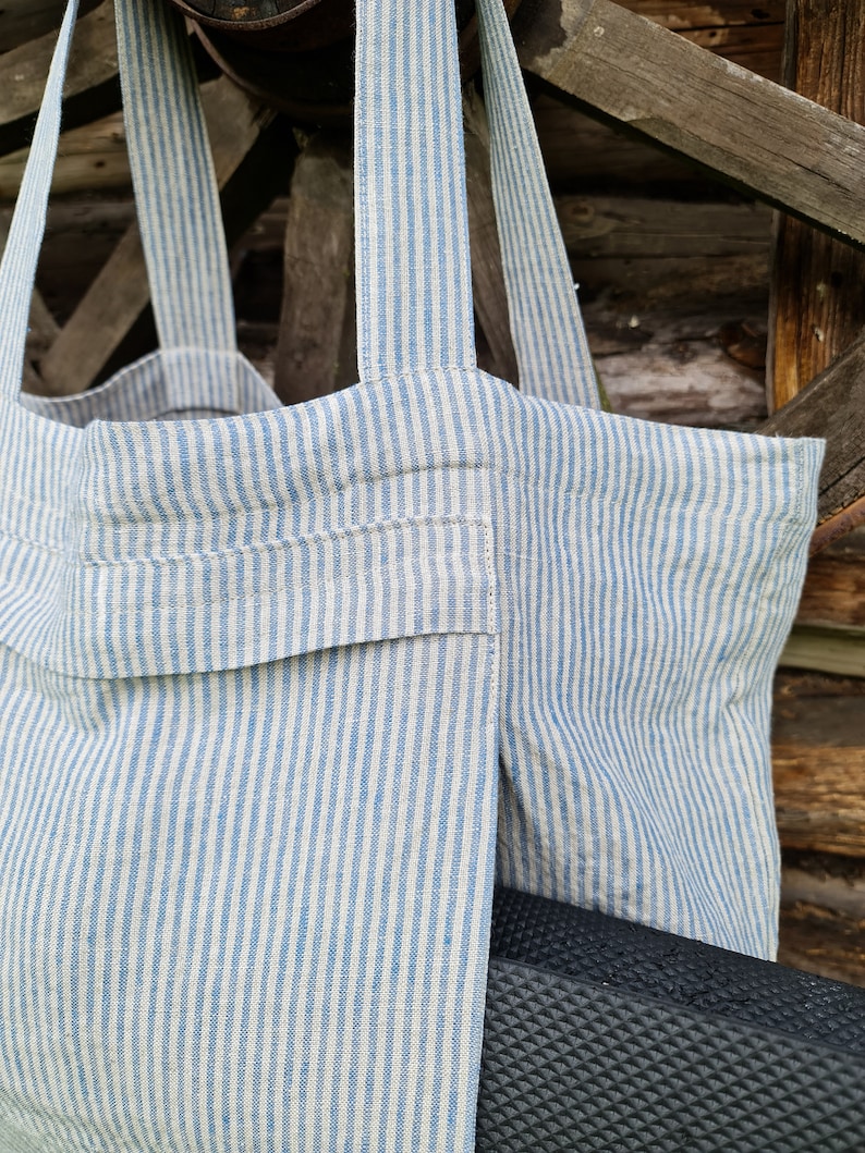May include: A striped tote bag made of natural linen fabric with blue stripes. The bag has a large pocket on the front and is hanging from a wooden structure.