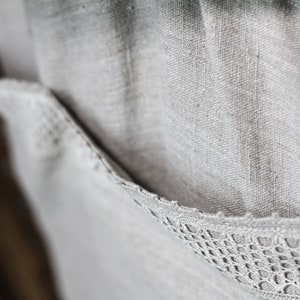 May include: A close-up of a light gray linen fabric with a delicate white lace trim. The fabric has a textured, woven appearance.