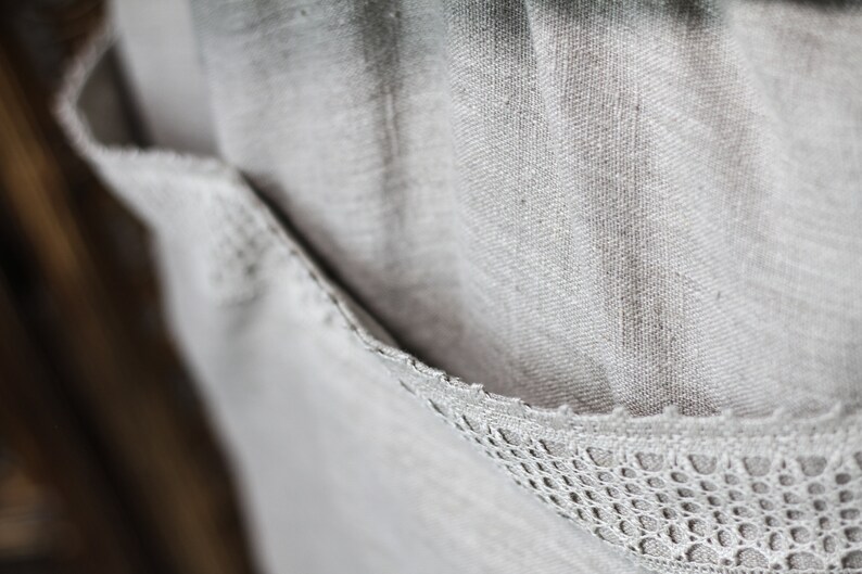 May include: A close-up of a light gray linen fabric with a delicate white lace trim. The fabric has a textured, woven appearance.