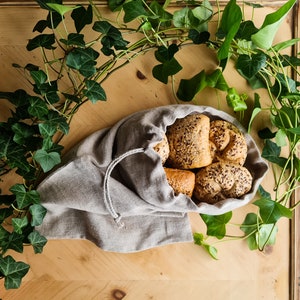 Op de afbeelding: Een lichtbruine linnen tas gevuld met zes vers gebakken broodjes. De tas ligt op een houten oppervlak met groene, bladerrijke wijnstokken eromheen.