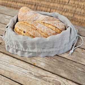 May include: Two loaves of bread in a gray linen bread bag. The bag is sitting on a wooden surface.