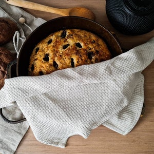 May include: A white kitchen towel draped over a cast iron pot with a loaf of bread inside. The bread is golden brown and has a few dark brown olives on top. There are two other loaves of bread in the background.