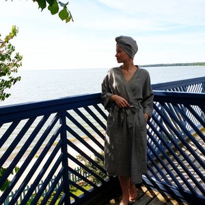 May include: A woman wearing a gray striped robe with a matching head wrap stands on a wooden deck overlooking a body of water. The deck has a blue railing with a diagonal pattern.