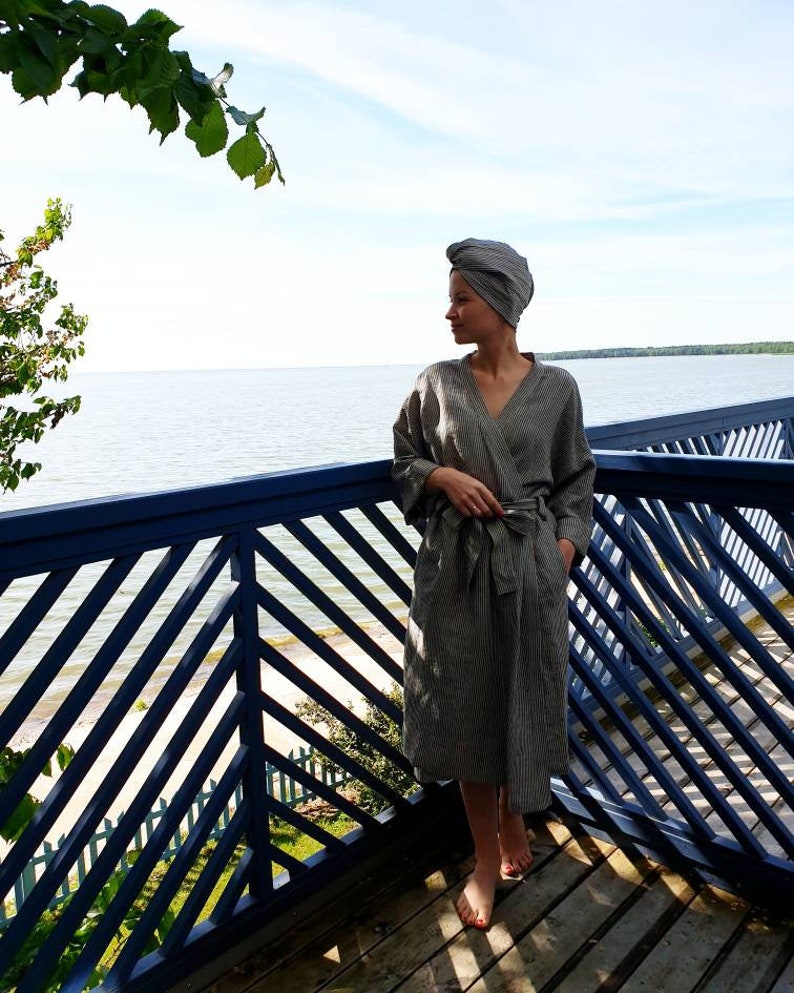May include: A woman wearing a gray striped robe with a matching head wrap stands on a wooden deck overlooking a body of water. The deck has a blue railing with a diagonal pattern.