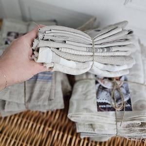 May include: A stack of natural linen tea towels, tied together with twine, on a woven wicker surface.