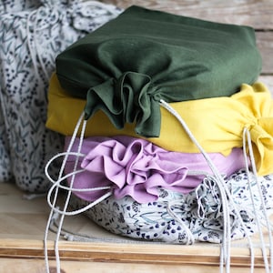 May include: A stack of four fabric drawstring bags in various colors and patterns. The top bag is dark green, the second is yellow, the third is purple, and the bottom bag is white with a floral pattern.