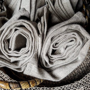 May include: A close-up of two rolls of natural linen fabric in a woven basket. The fabric is a light beige color and has a rough texture.