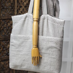 May include: A large gray linen tote bag with white lace trim. The bag is being held by a wooden hand.