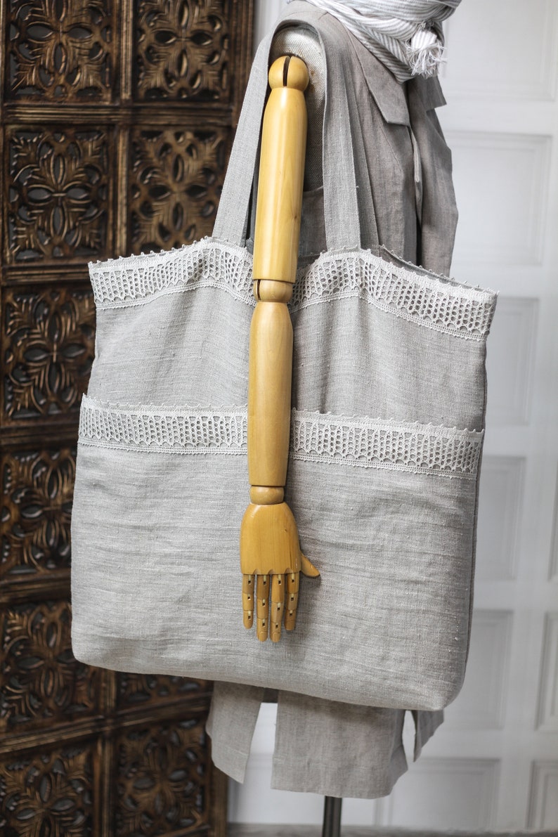May include: A large gray linen tote bag with white lace trim. The bag is being held by a wooden hand.
