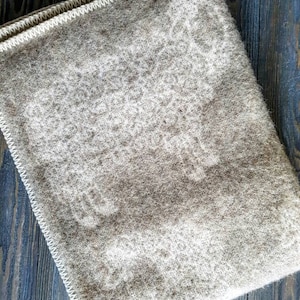 May include: A beige wool blanket with a subtle woven pattern. The blanket has a white border and is folded in half.