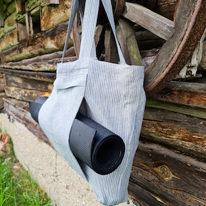 May include: A blue and white striped tote bag with a black yoga mat tucked inside. The bag is hanging on a wooden wall.