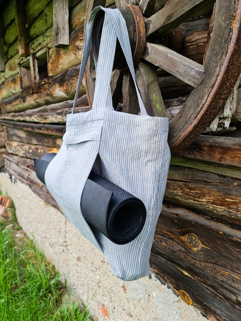 May include: A blue and white striped tote bag with a black yoga mat tucked inside. The bag is hanging on a wooden wall.