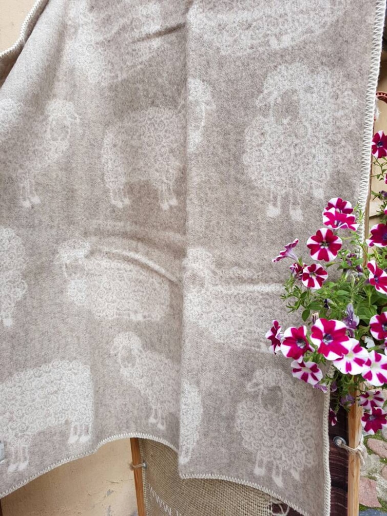 May include: A beige wool throw blanket with a white sheep pattern. The blanket is folded and draped over a wooden chair. A potted plant with pink and white flowers is in the background.
