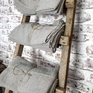 May include: A stack of striped linen fabric bundles tied with twine, displayed on a rustic wooden ladder against a white brick wall.