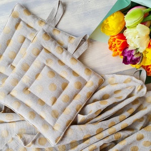 Puede incluir: Dos agarraderas de olla beige con lunares dorados y un diseño acolchado. Las agarraderas están sobre una tela beige con lunares dorados.