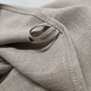 May include: A beige linen textile featuring a visible woven texture and a small fabric loop tag. The tag is printed with the text The Raw Linen and includes a small black circular logo icon. The fabric has a hemmed edge with visible stitching.