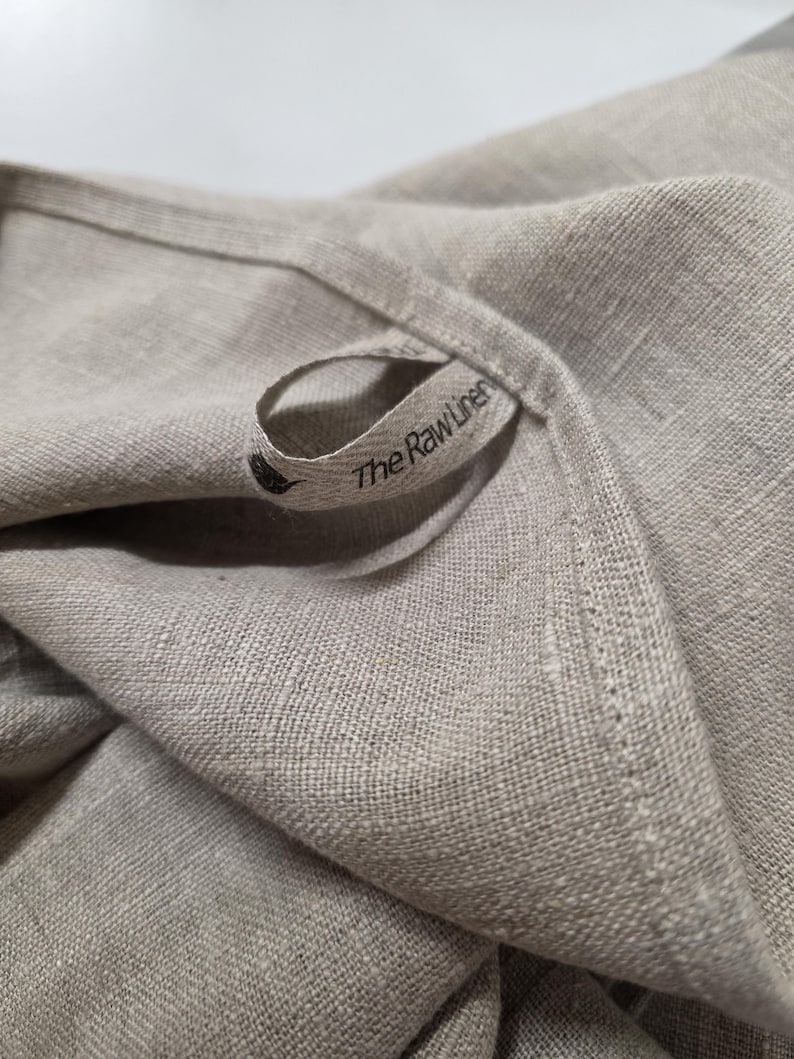 May include: A beige linen textile featuring a visible woven texture and a small fabric loop tag. The tag is printed with the text The Raw Linen and includes a small black circular logo icon. The fabric has a hemmed edge with visible stitching.