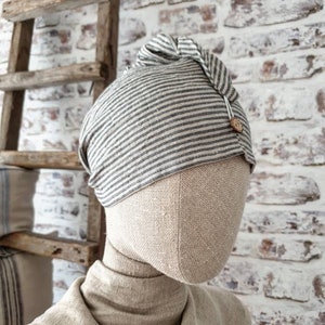May include: A gray and white striped linen beanie with a knot on top and a small wooden button. The beanie is on a fabric-covered mannequin head.