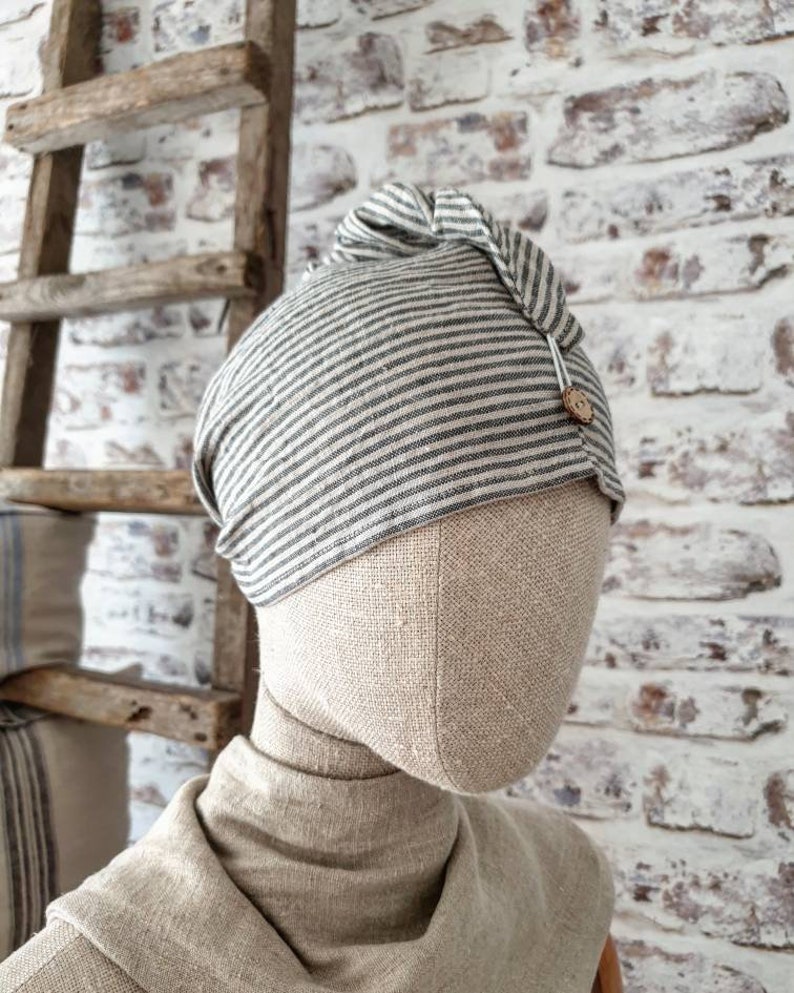 May include: A gray and white striped linen beanie with a knot on top and a small wooden button. The beanie is on a fabric-covered mannequin head.