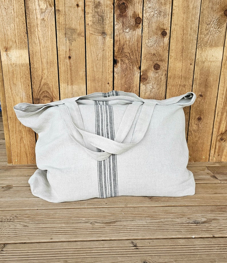 May include: A light gray linen tote bag with a vertical stripe design in the center. The bag has two handles and is sitting on a wooden surface. The stripe design is composed of gray and white stripes.