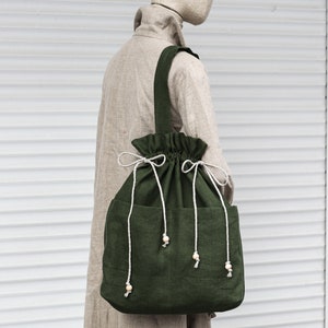 May include: A large, green linen tote bag with a drawstring closure and wooden bead accents. The bag has two pockets on the front and is worn by a mannequin.