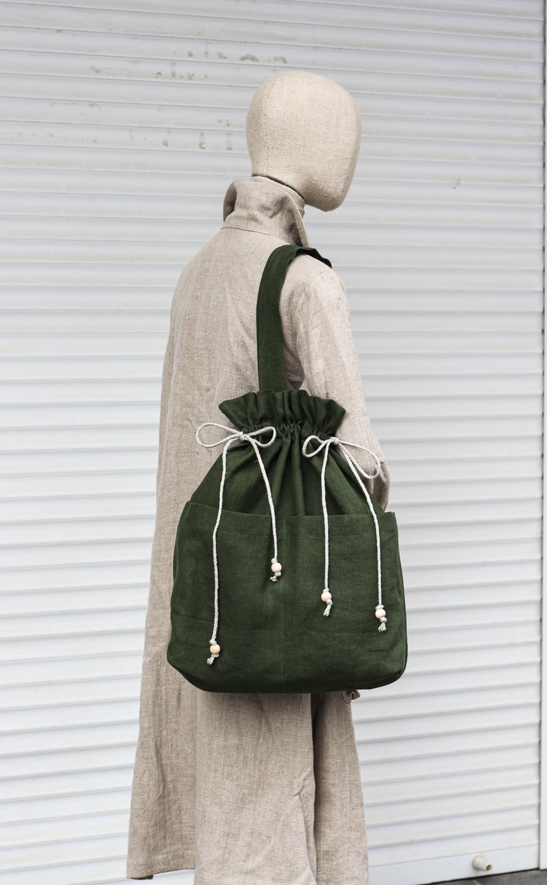 May include: A large, green linen tote bag with a drawstring closure and wooden bead accents. The bag has two pockets on the front and is worn by a mannequin.