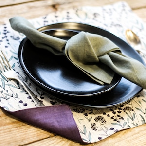 May include: A black dinner plate with a smaller black plate on top. A green linen napkin is tied in a knot on top of the plates. The plates are on a white and green floral placemat.