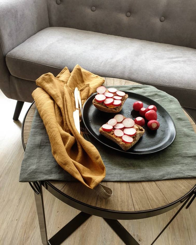 Puede incluir: Dos s&aacute;ndwiches abiertos con rodajas de r&aacute;bano encima, servidos en un plato negro con un acompa&ntilde;amiento de r&aacute;banos rojos. El plato est&aacute; sobre una mesa de madera con una servilleta de lino gris y amarillo mostaza.