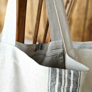 May include: A light-colored tote bag with a striped design on the front. The bag has long straps and a zipper closure. The bag is made of a textured fabric and is hanging on a wooden chair.