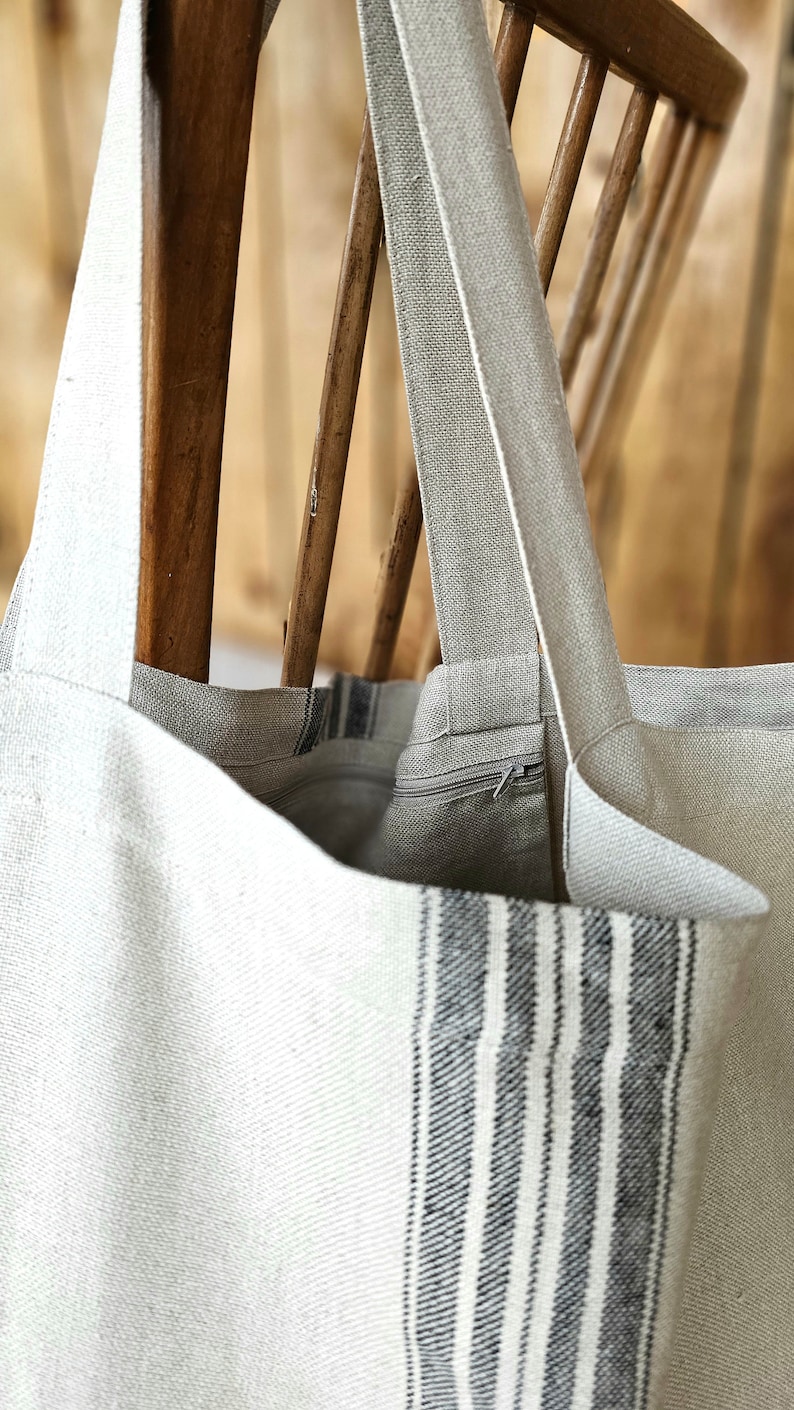 May include: A light-colored tote bag with a striped design on the front. The bag has long straps and a zipper closure. The bag is made of a textured fabric and is hanging on a wooden chair.