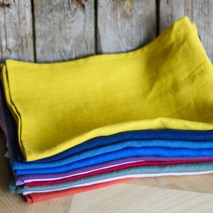 Puede incluir: Una pila de diez toallas de t&eacute; de lino en varios colores, incluyendo amarillo, azul, morado, rojo, verde y naranja. Las toallas est&aacute;n dobladas y apiladas sobre una superficie de madera.