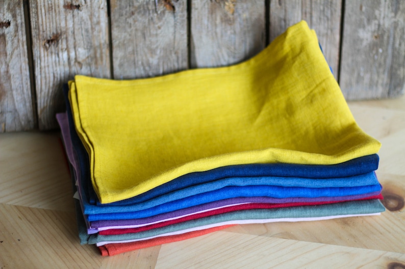Puede incluir: Una pila de diez toallas de t&eacute; de lino en varios colores, incluyendo amarillo, azul, morado, rojo, verde y naranja. Las toallas est&aacute;n dobladas y apiladas sobre una superficie de madera.