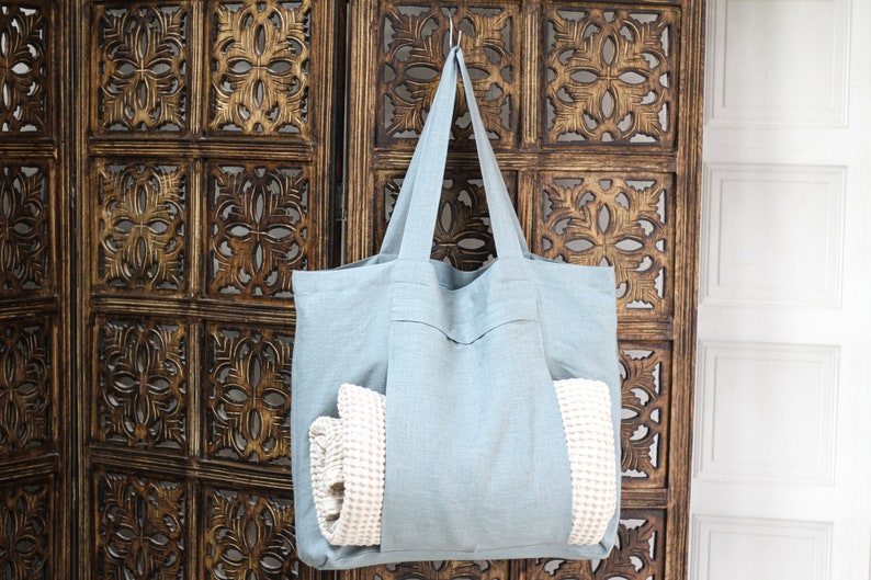 May include: A light blue tote bag made of linen with a white waffle weave towel inside. The bag is hanging on a wooden screen with intricate carvings.
