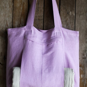 May include: A lavender linen tote bag with a large pocket on the front. A white crocheted towel is tucked into the pocket.