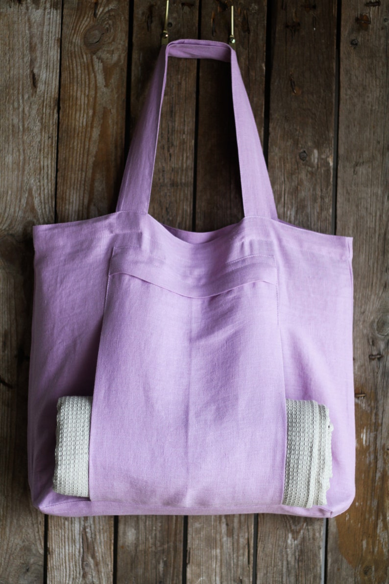 May include: A lavender linen tote bag with a large pocket on the front. A white crocheted towel is tucked into the pocket.