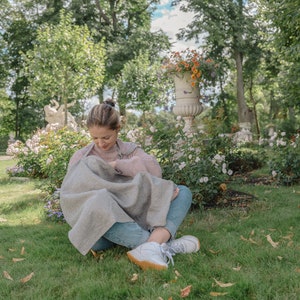 Pu&ograve; includere: Una donna &egrave; seduta sull'erba in un parco, vestita con un maglione rosa chiaro e jeans blu. Tiene in braccio un bambino avvolto in una coperta grigia e bianca. La donna sta allattando il bambino.