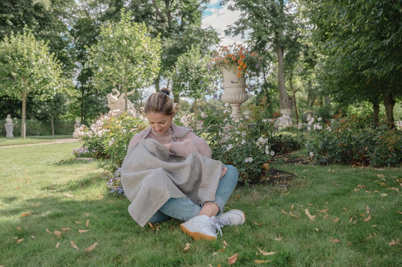 Pu&ograve; includere: Una donna &egrave; seduta sull'erba in un parco, vestita con un maglione rosa chiaro e jeans blu. Tiene in braccio un bambino avvolto in una coperta grigia e bianca. La donna sta allattando il bambino.