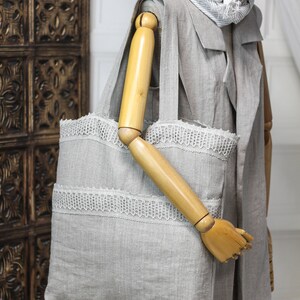 May include: A gray linen tote bag with white lace trim. The bag has a wooden handle and is shown on a mannequin.