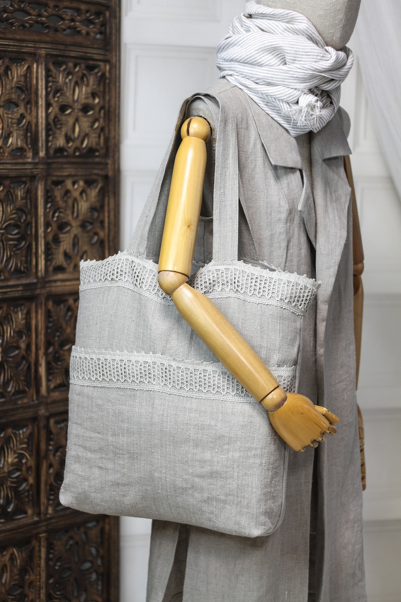 May include: A gray linen tote bag with white lace trim. The bag has a wooden handle and is shown on a mannequin.
