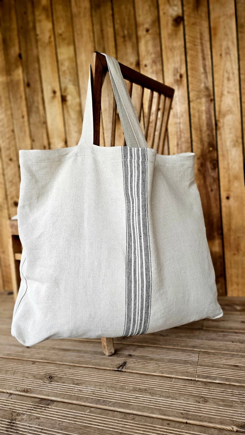 May include: A large, light gray tote bag with a vertical stripe design in black and white. The bag has long handles and is made of a textured fabric. It is resting on a wooden chair and a wooden surface.