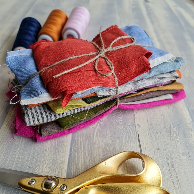 May include: A stack of colorful fabric scraps tied together with twine. The fabric scraps are in various shades of blue, red, green, yellow, and brown. There are three spools of thread in the background, one orange, one blue, and one pink.