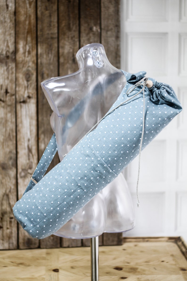 May include: A blue yoga mat bag with white polka dots and a drawstring closure. The bag has a long strap for carrying over the shoulder.