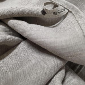 May include: A light beige, textured fabric cloth featuring a small, folded fabric loop tag. The tag displays a small black icon of a thistle and the printed text "The Raw Linen" in a simple, dark, sans-serif font, stitched onto the edge.
