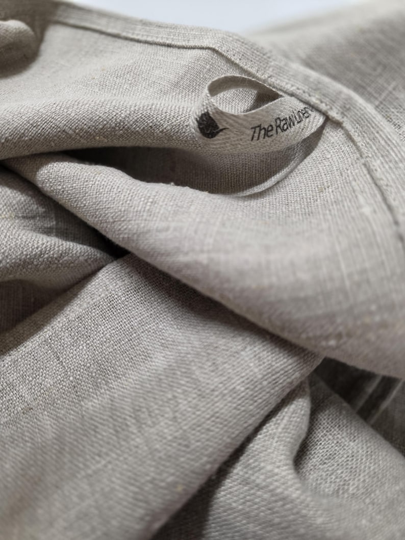 May include: A light beige, textured fabric cloth featuring a small, folded fabric loop tag. The tag displays a small black icon of a thistle and the printed text "The Raw Linen" in a simple, dark, sans-serif font, stitched onto the edge.