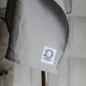 Pu&ograve; includere: Un fasciatoio per neonati in lino grigio con un'etichetta bianca che dice "Buddy Buddy Nest by The Raw Linen".