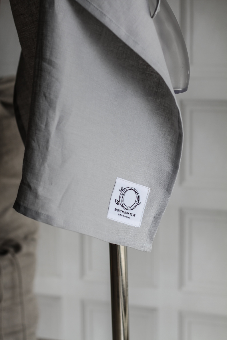 Pu&ograve; includere: Un fasciatoio per neonati in lino grigio con un'etichetta bianca che dice "Buddy Buddy Nest by The Raw Linen".
