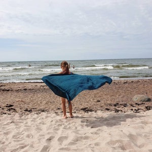 May include: A young person standing on a sandy beach with a teal blue scarf flowing in the wind. The ocean is in the background.