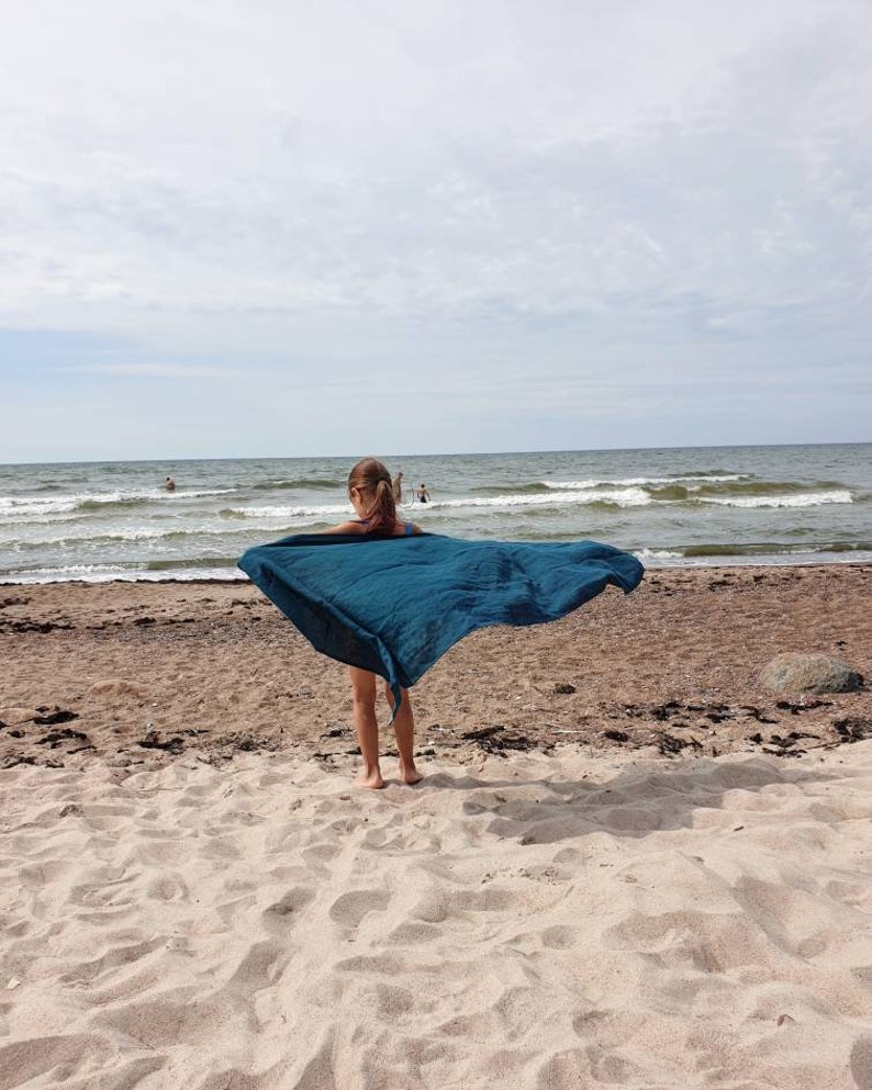 May include: A young person standing on a sandy beach with a teal blue scarf flowing in the wind. The ocean is in the background.