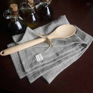 May include: A light brown wooden spoon tied with a jute twine bow, resting on a folded grey linen kitchen towel. The towel features a small white fabric tag with the text "HANDMADE IN LITHUANIA". Three glass cruets with cork stoppers are visible.